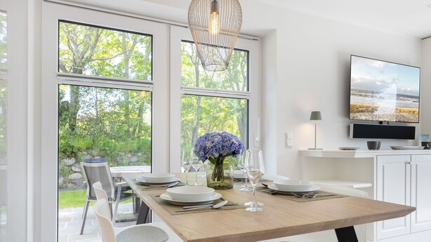 A modern dining area with a wooden table set for four, large windows offering a view of greenery, and a flat-screen TV mounted on the wall.