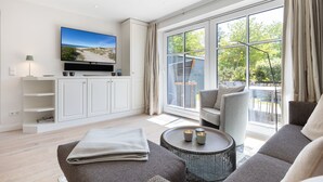 A modern living room with a flat-screen TV, a sofa, a coffee table, and a view of the outdoors through large windows.