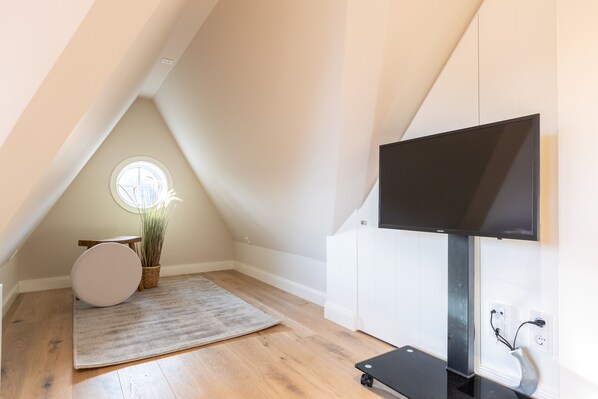 A modern, well-lit attic room with a large TV, a round mirror, and a small table with a plant. - Das Fröddenhues Befindet Sich in Sehr Ruhiger und Geschützter Lage (Sylt)