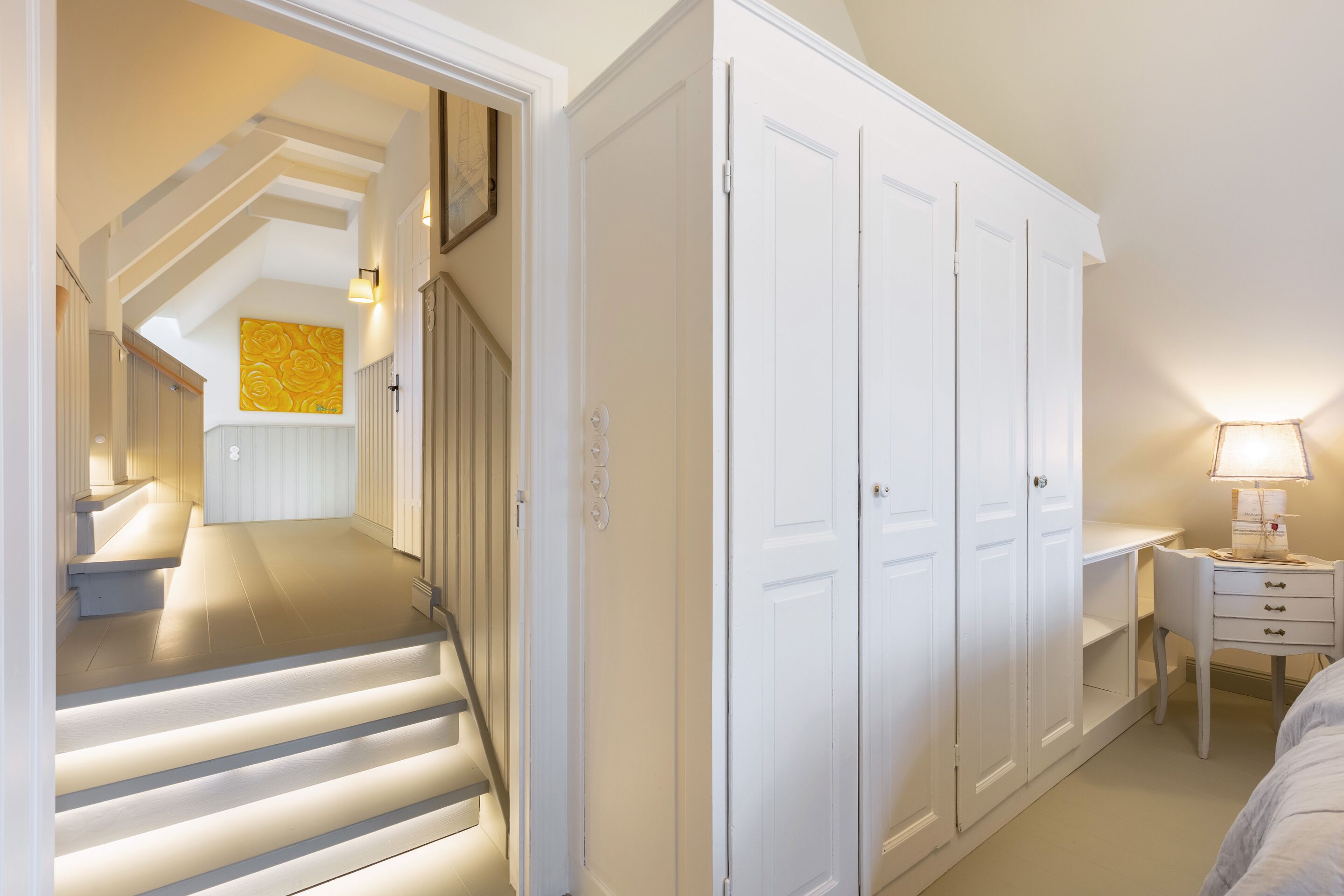 A bedroom with a bed, a desk with a lamp, a wardrobe, and a staircase leading to another room.