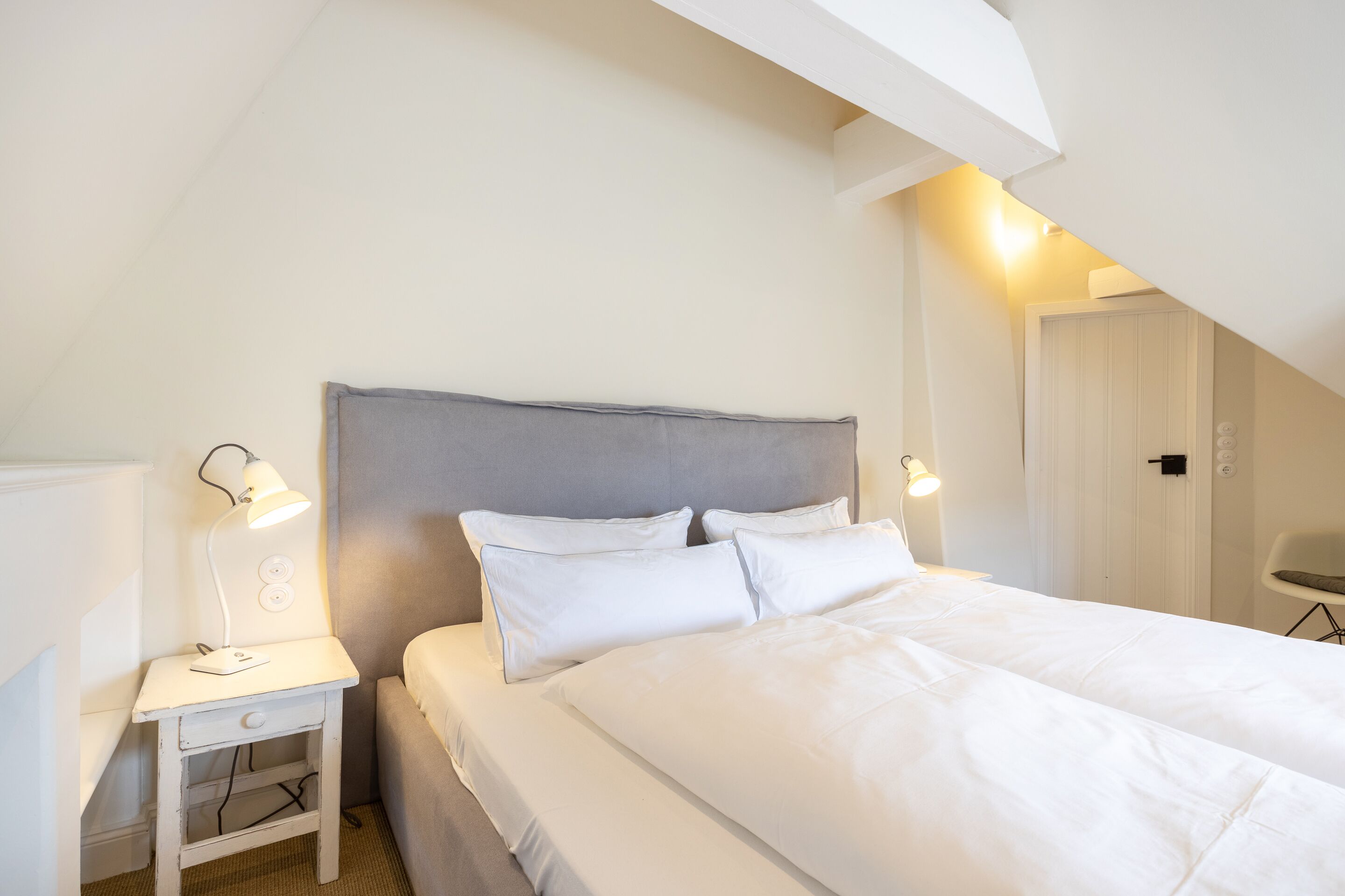 A neatly made bed in a room with a sloped ceiling, a bedside table with a lamp, and a chair.
