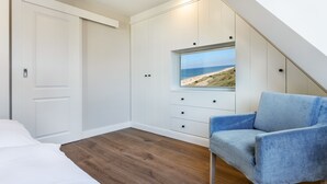 Ein Schlafzimmer mit Holzboden, einem Bett, einem blauen Sessel, einem Kleiderschrank und einem Fenster mit Blick auf den Strand.