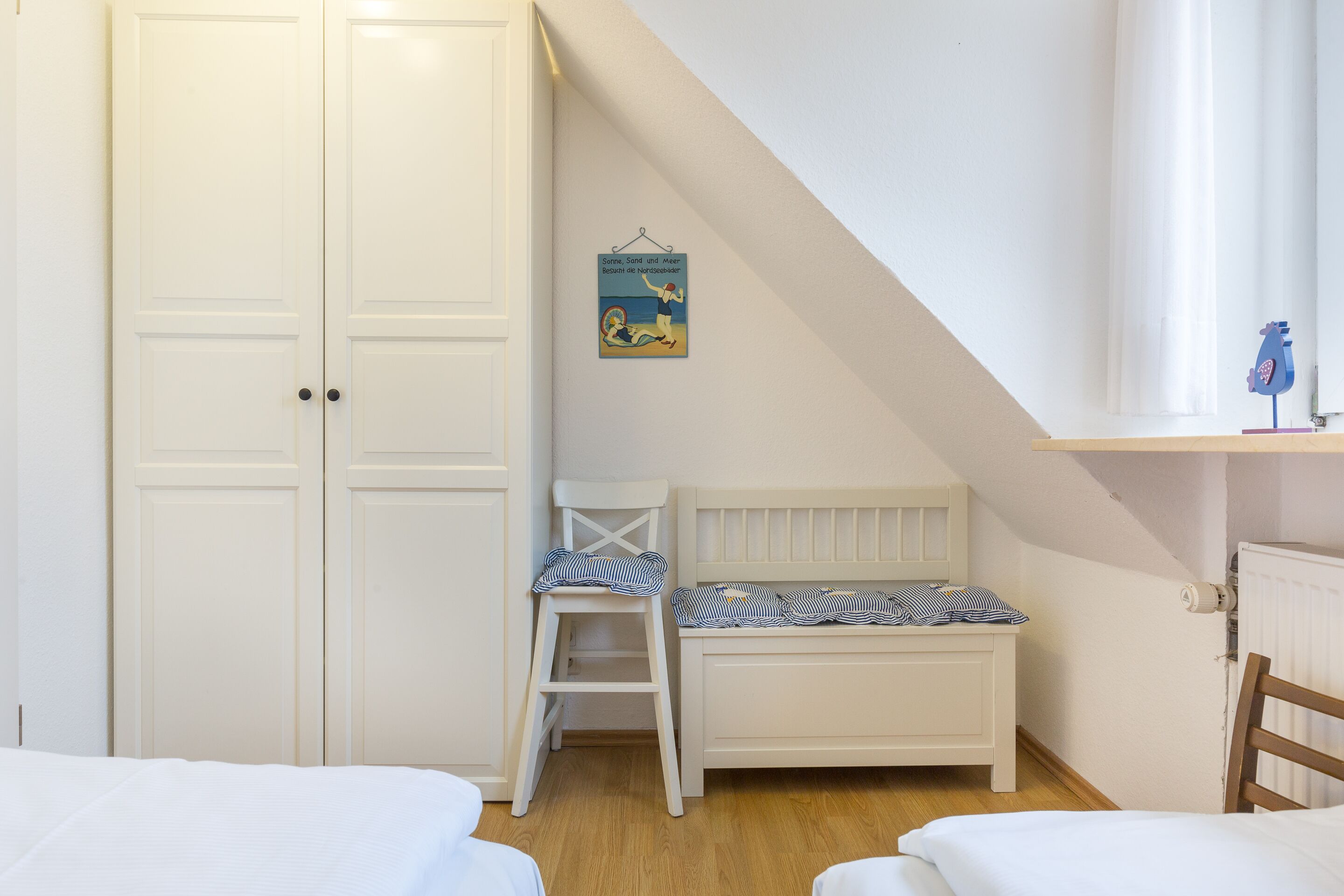 A bedroom with a white bed, a small white chair, a white wardrobe, and a poster on the wall.