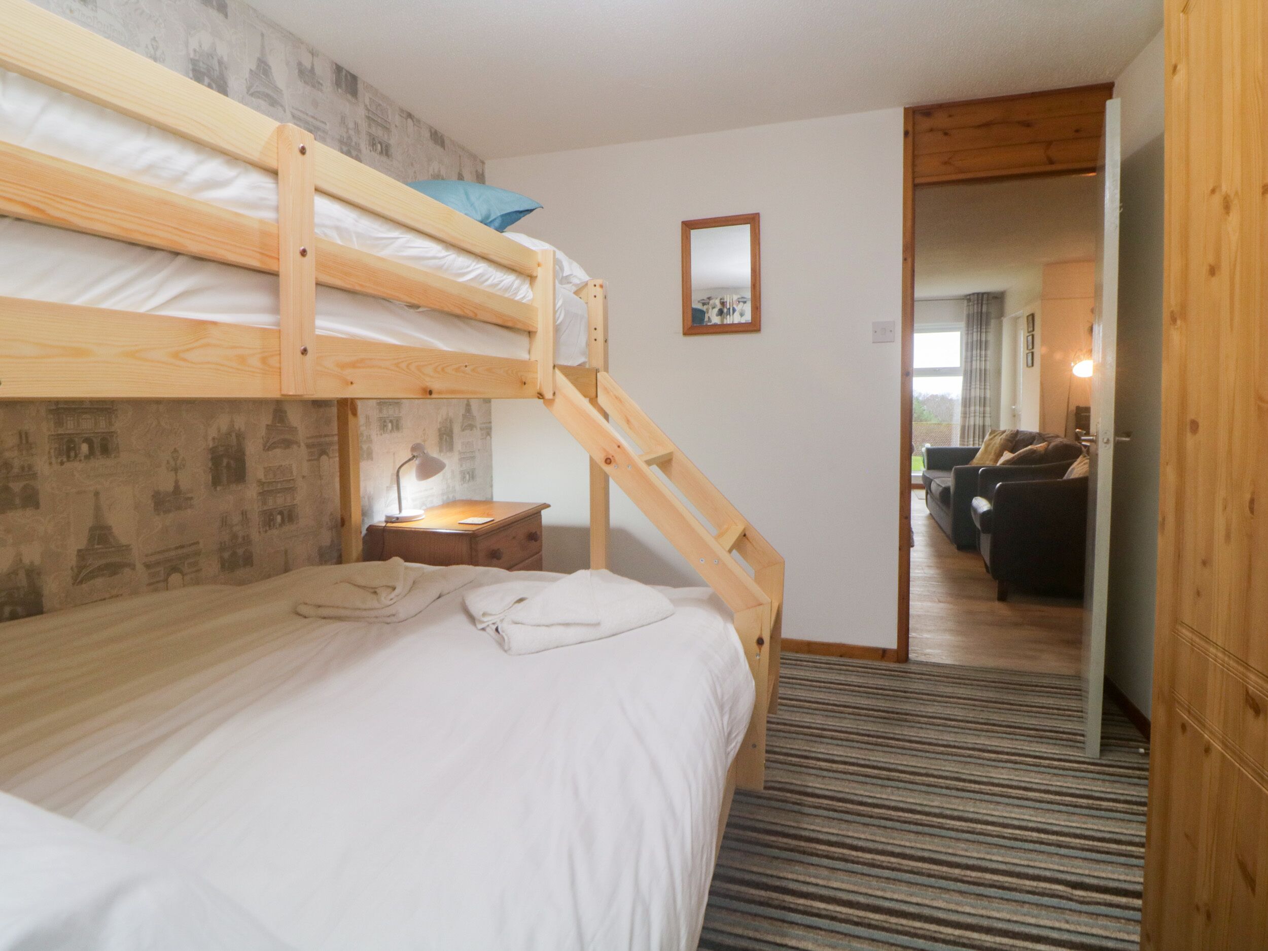 A bunk bed room with a desk and lamp, a mirror on the wall, and a view into a living area with a sofa.
