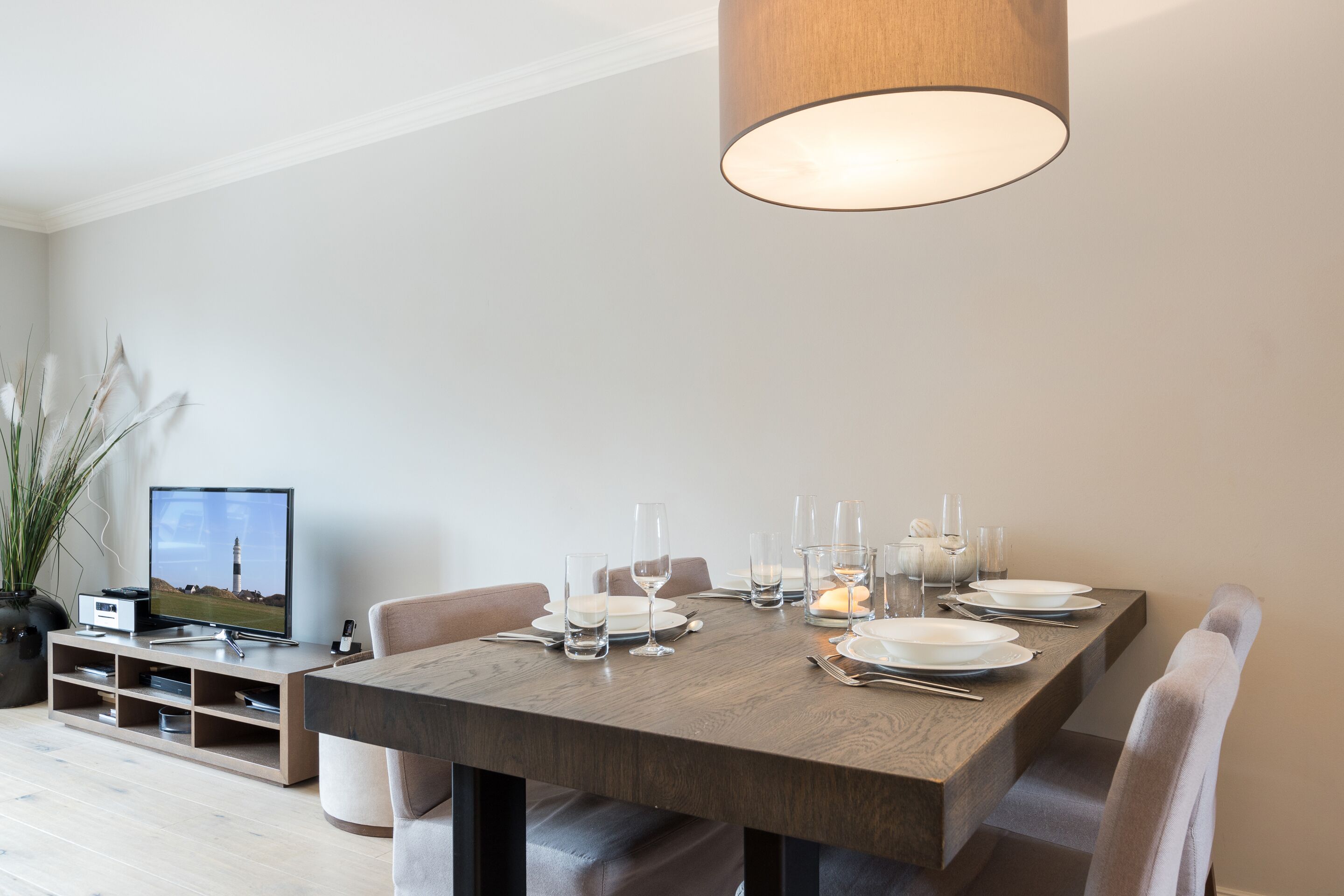 Un comedor con una mesa de madera preparada para cuatro, un televisor sobre un soporte y una planta en maceta.