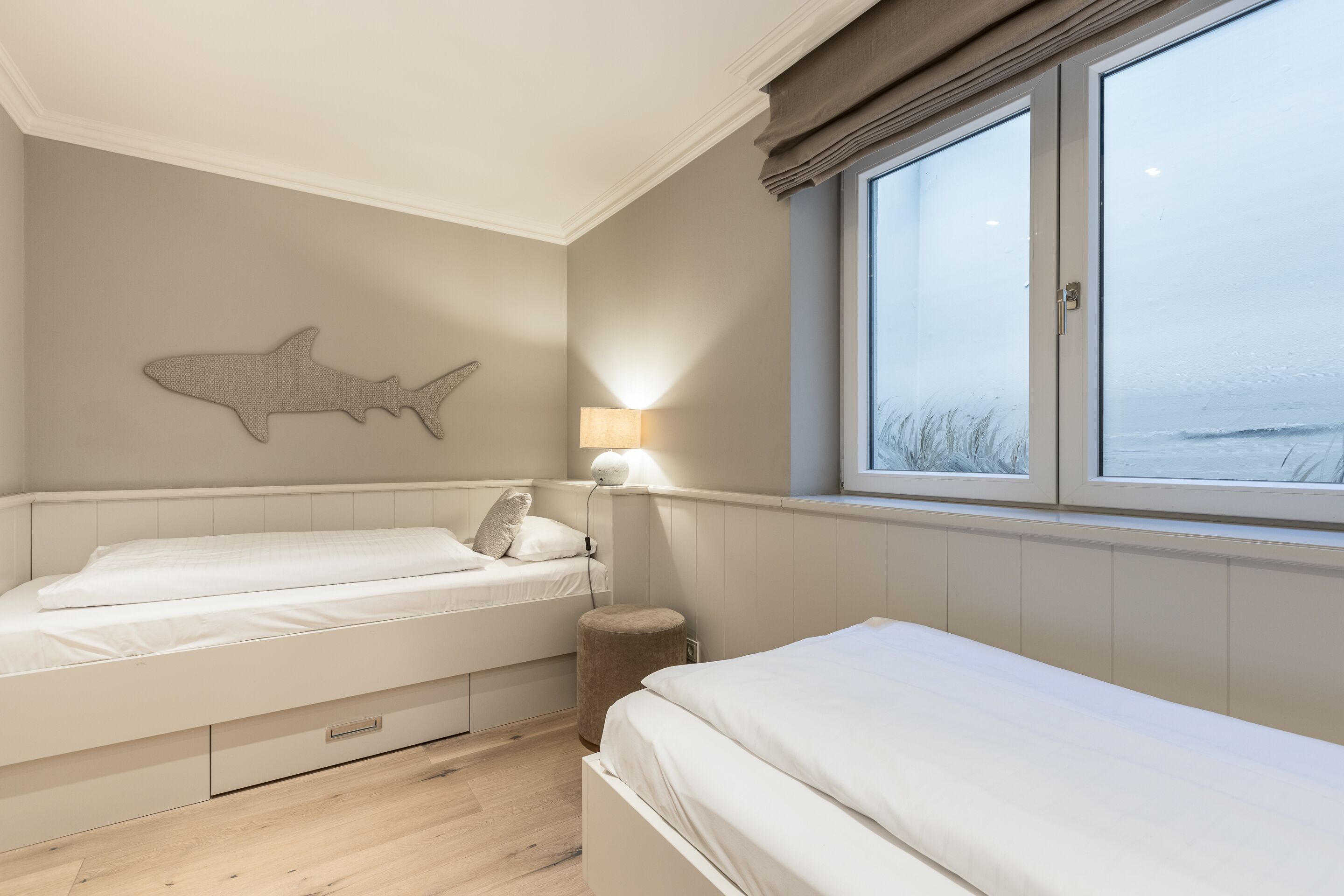 A bedroom with two beds, a wall-mounted shark decoration, a bedside lamp, and a window with a view of the sea.