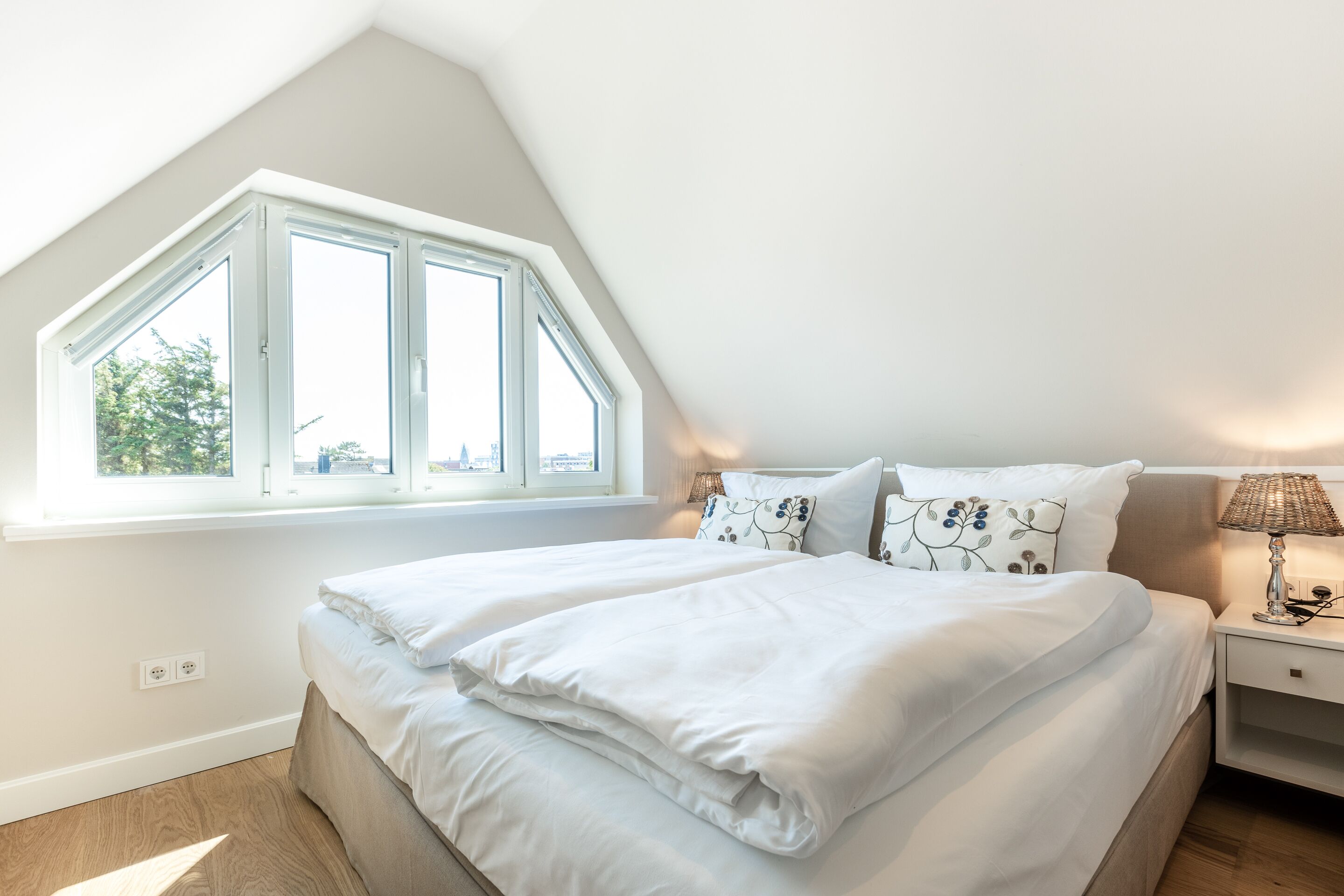 A bedroom with a large window, a bed with white linens, a bedside lamp, and a nightstand.