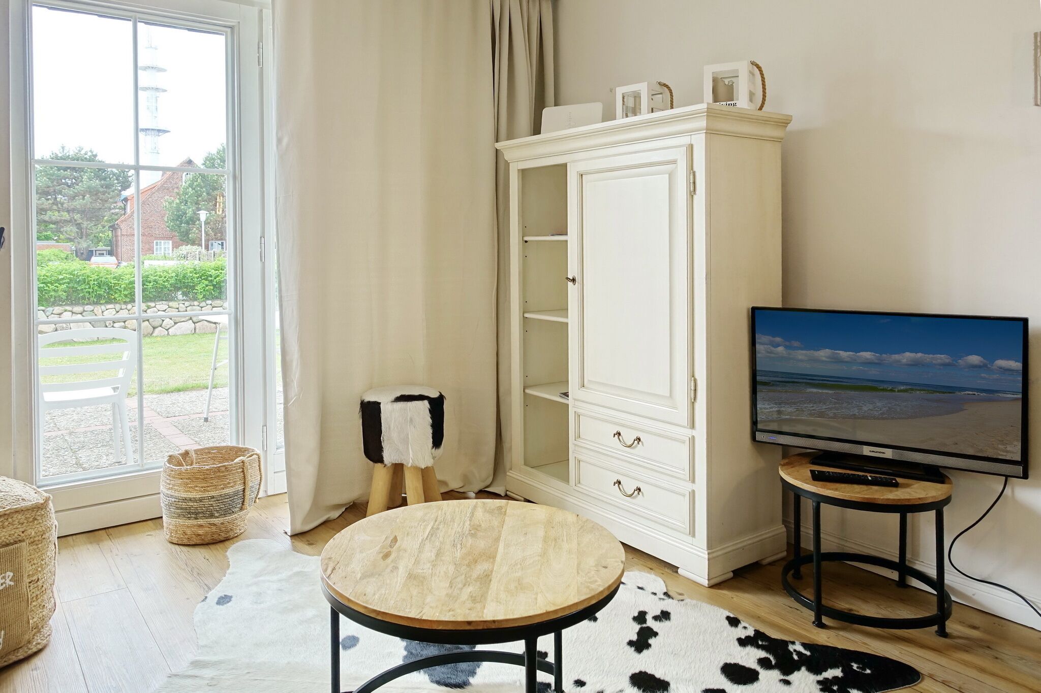 Ett rum med ett stort fönster, ett vitt skåp, ett runt träbord, en tv på ett stativ och en matta av kohud.