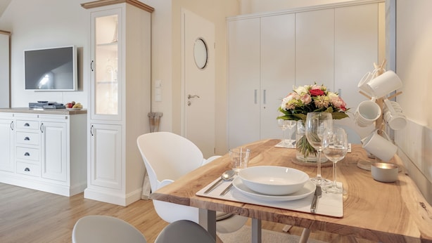 A dining area with a wooden table set for four, white chairs, a TV, and a cabinet with a mirror. - Das Appartement Senskin für 2 Personen in Ruhiger Lage in Westerland (Sylt)