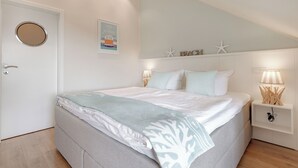 A bedroom with a bed, nightstands, a wall-mounted shelf with decorative items, and a door with a circular mirror. - Das Appartement Senskin für 2 Personen in Ruhiger Lage in Westerland (Sylt)