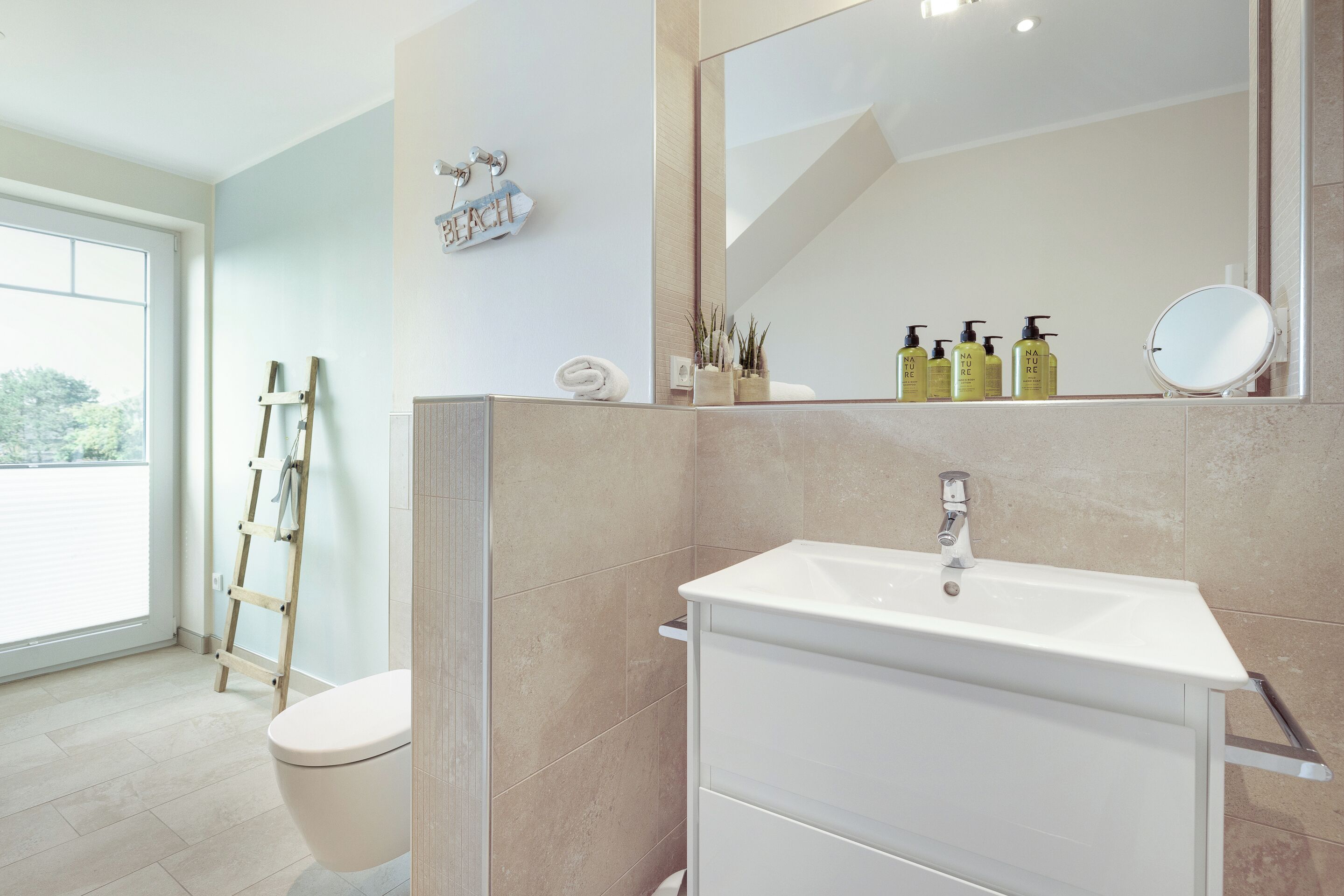 Une salle de bain avec un lavabo blanc, des carreaux de couleur beige, un porte-serviettes et une échelle appuyée contre le mur.