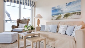 A living room with a beige sofa, a wicker chair, a small table with a lamp, and a large window with a view of the beach.