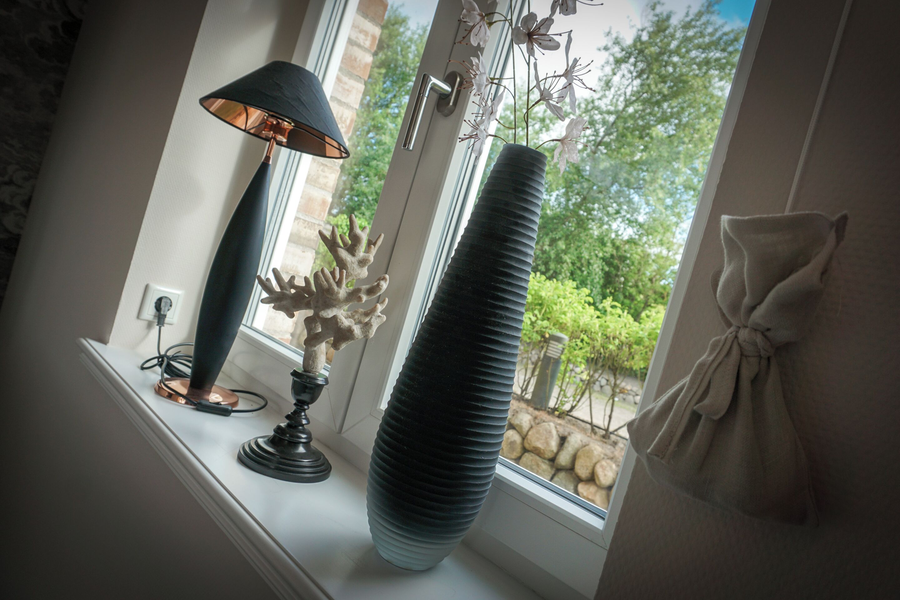 A lamp, a vase, and a window with a view of trees and a stone wall.