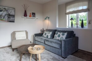 A living room with a grey sofa, a round coffee table, a beige armchair, and a window with curtains.