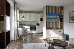 Cozinha moderna com armários brancos, sala de jantar com mesa redonda e cadeiras, e sala de estar com sofá e mesa de centro.