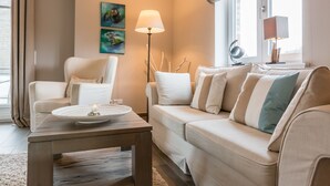 Een moderne woonkamer met een beige bank, een witte fauteuil, een houten salontafel en een vloerlamp. Twee abstracte schilderijen hangen aan de muur en er is een raam met uitzicht op het omliggende landschap.
