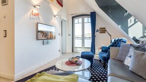 Ein modernes Wohnzimmer mit einem Glastisch, einer Couch und einem blauen Sessel. Es gibt ein großes Fenster mit Vorhängen, ein Bild an der Wand und eine Decke mit einem Oberlicht.