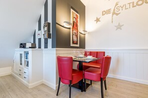 Uma sala de jantar com uma mesa posta para quatro pessoas, cadeiras vermelhas, uma luminária fixada na parede e um cartaz.
