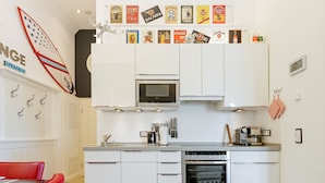 Eine moderne Küche mit weißen Schränken, einer Mikrowelle, einem Backofen und einer Kaffeemaschine. An der Wand hängen verschiedene Küchenutensilien, und ein Surfbrett dient als Dekoration.