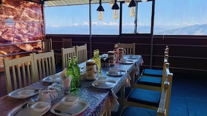 A dining area with a table set for four, chairs, and a view of snow-capped mountains through large windows. - Family in the Hills by Nature's Glamping (Kosiyakutoli)