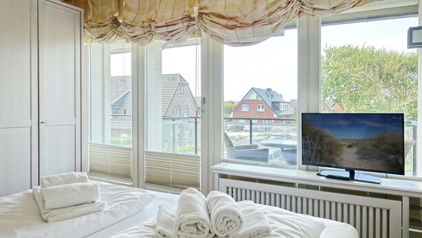 Et soveværelse med seng, et vindue med persienner, et fjernsyn på et stativ og en radiator.