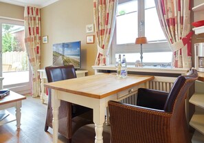 A dining area with a wooden table, wicker chairs, and a view of the outdoors through a window with curtains. - 2-raum Appartement mit ca. 45m ² Nutzfläche im Erd- und Untergeschoss (Wenningstedt-Braderup (Sylt))