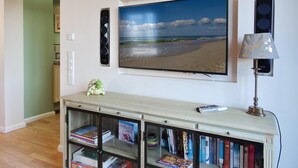 Una sala de estar con un televisor de pantalla plana grande montado en la pared, una estantería llena de libros, una lámpara y un pequeño objeto decorativo sobre una consola.