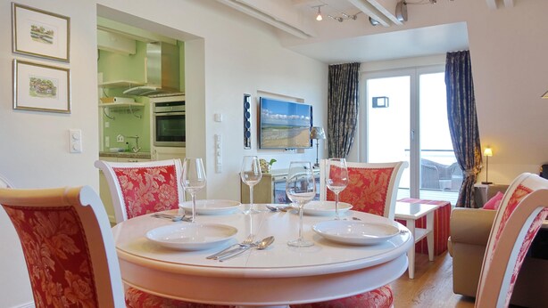Comedor con una mesa redonda preparada para cuatro personas, rodeada de sillas tapizadas rojas. Se ve una cocina al fondo, y un ventanal con cortinas a la derecha.