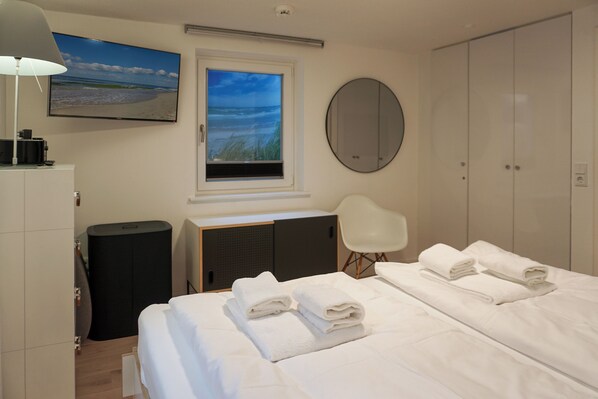 A hotel room with a bed, a TV, a window with a beach view, a mirror, and a wardrobe. - 2-raum-appartement im Erd- und UG mit ca. 53m² Nutzfläche für Max. Zwei Personen (Wenningstedt-Braderup (Sylt))