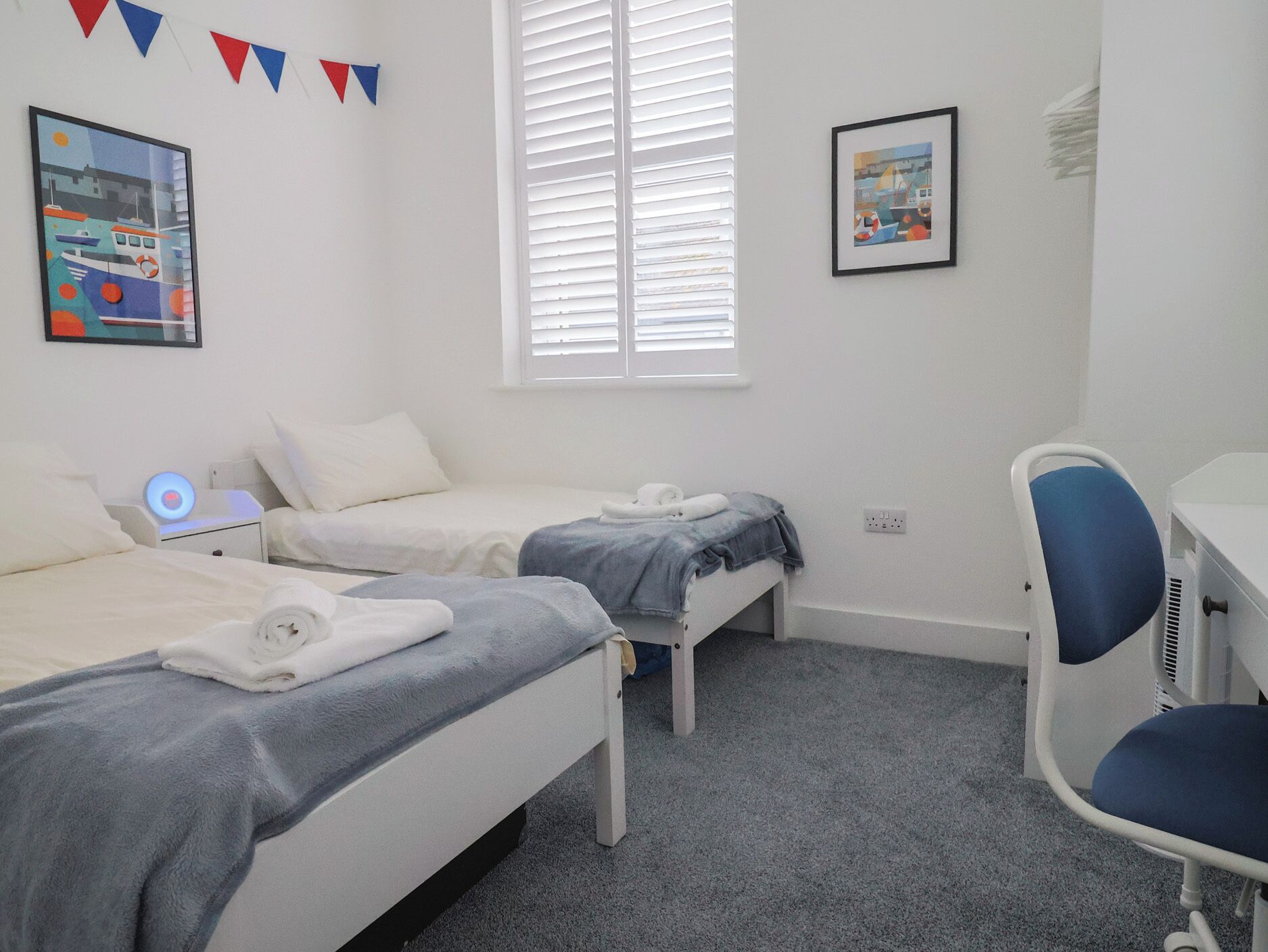 A bedroom with two beds, a desk, and a chair. There are two framed pictures on the wall.