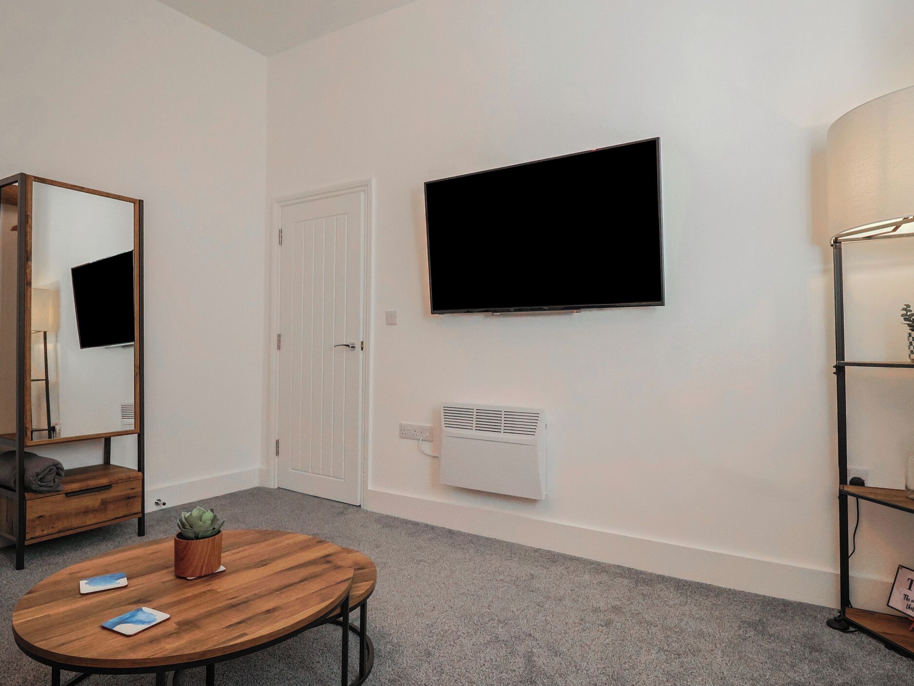 A room with a round wooden table, a mounted flat-screen TV, a white radiator, a white door, and a mirror on the left wall.