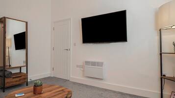 A room with a round wooden table, a mounted flat-screen TV, a white radiator, a white door, and a mirror on the left wall.