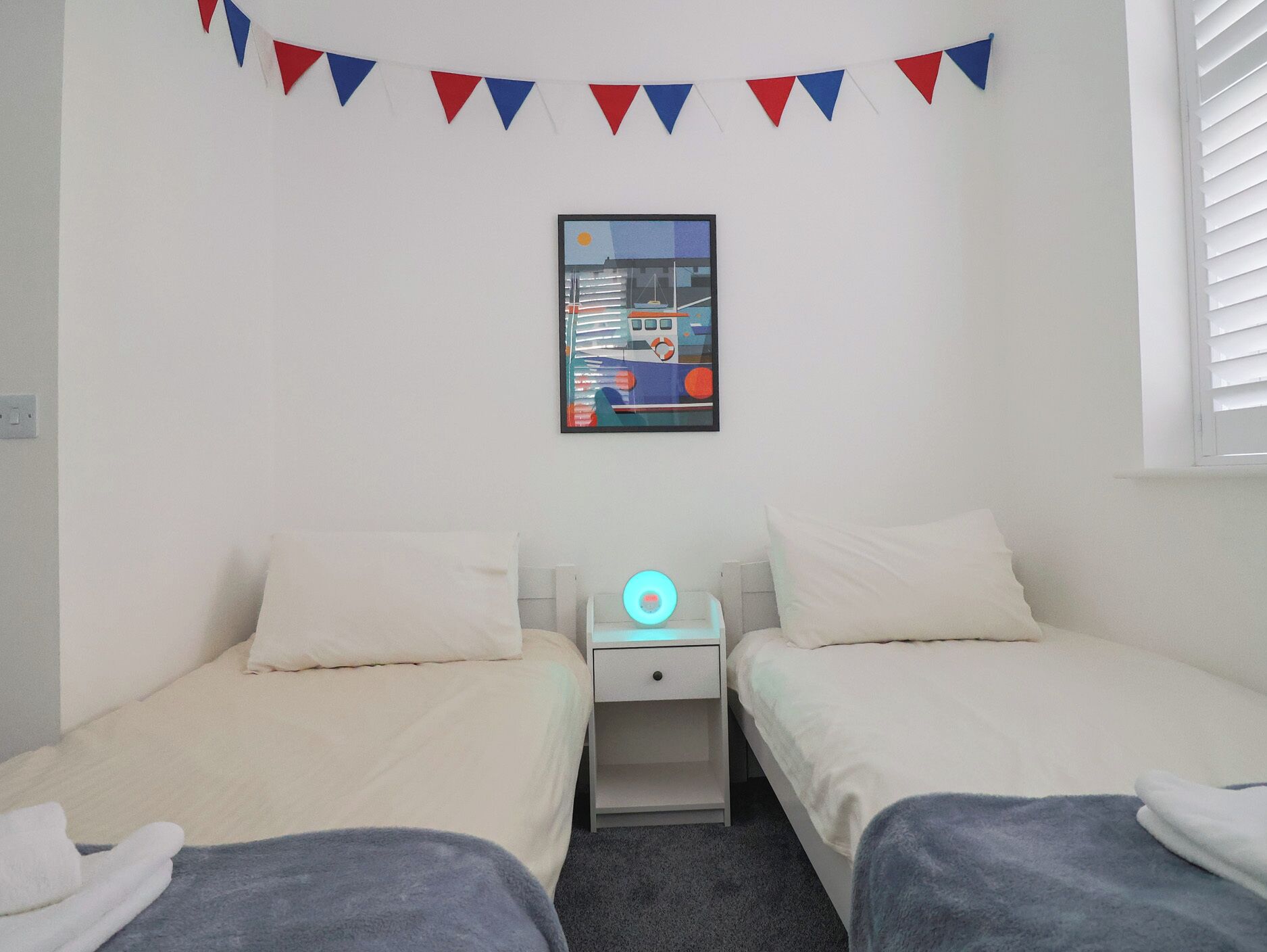 A bedroom with two beds, a nightstand, a wall-mounted picture, and a red and blue bunting decoration.