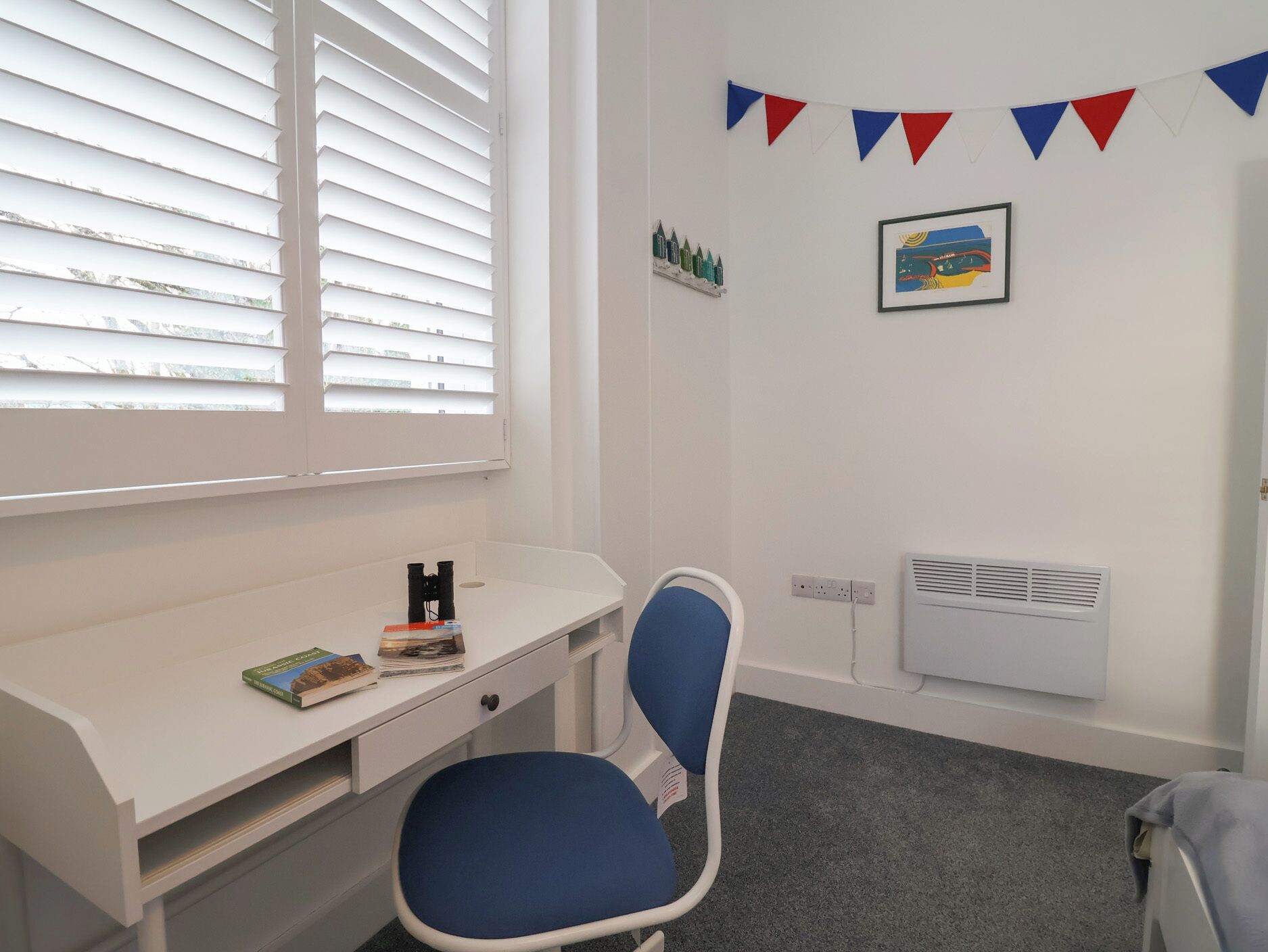 Ein Zimmer mit weißem Schreibtisch, blauem Stuhl, Heizkörper und einem Fenster mit Jalousien.