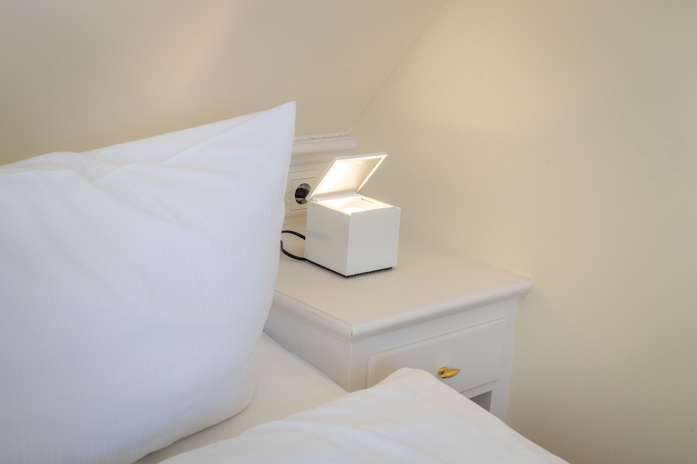 En pænt redt seng med hvide sengetøj og puder, et natbord med en hvid lampe, og en lille skuffe med en gylden knap.