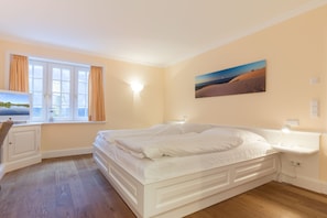 Un dormitorio con una cama grande, una ventana con cortinas, una mesita de noche con lámpara y un cuadro colgado en la pared.