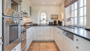 A modern kitchen with white cabinetry, a large island, stainless steel appliances, and a view of the outdoors through a window. - Strand, Meer & Luxus (Wenningstedt-Braderup (Sylt))