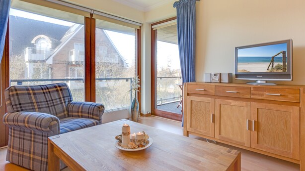 Ein Zimmer mit einer Couch, einem Tisch mit Kerzen, einem Fernseher und Blick auf den Strand durch große Fenster.