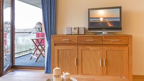 Ein Zimmer mit einem Holzkasten, einem Fernseher, der einen Sonnenuntergang über dem Meer zeigt, und einem Balkon mit Tisch und Stühlen.