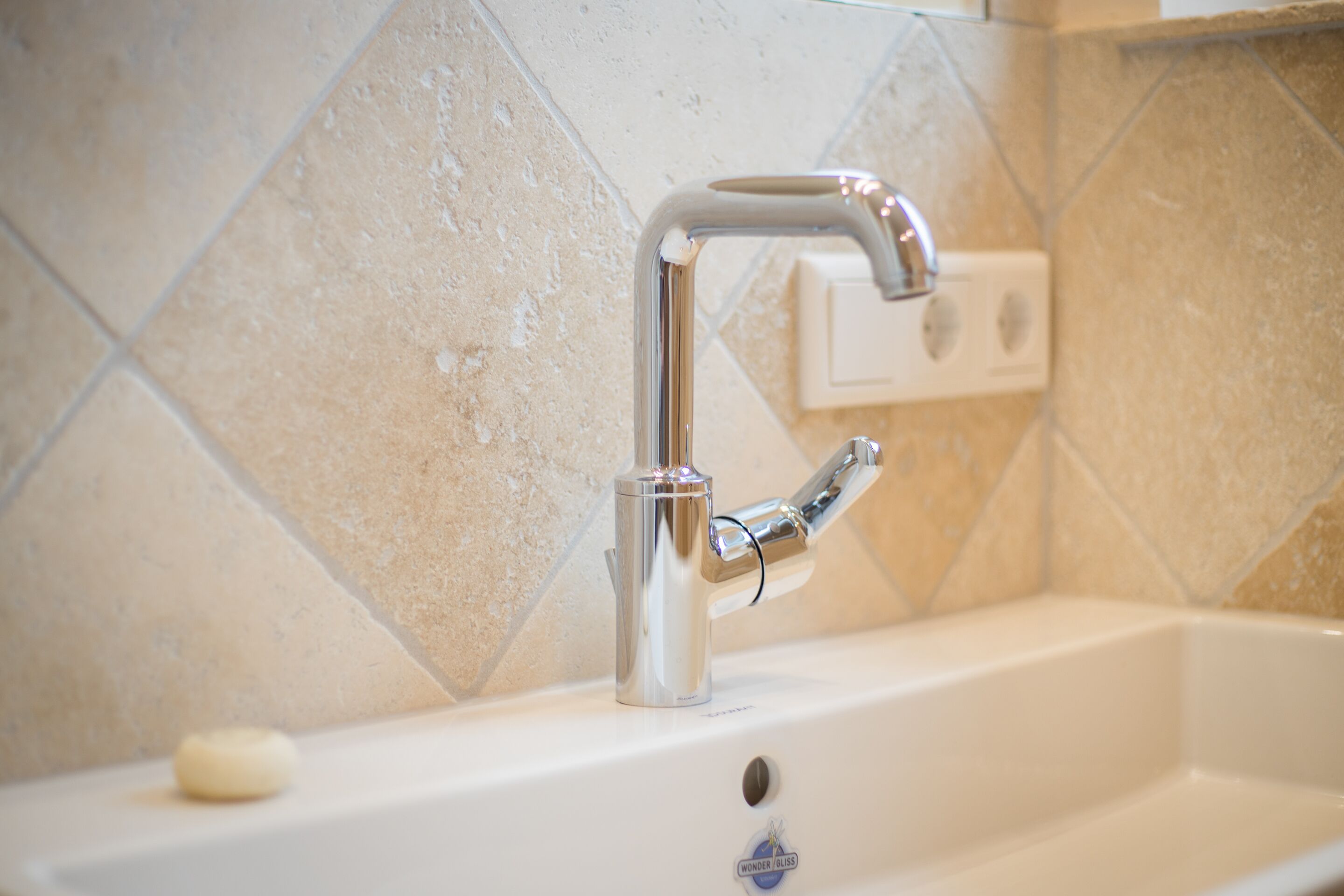 A bathroom sink with a chrome faucet, a soap dish, and a white wall with beige tiles.