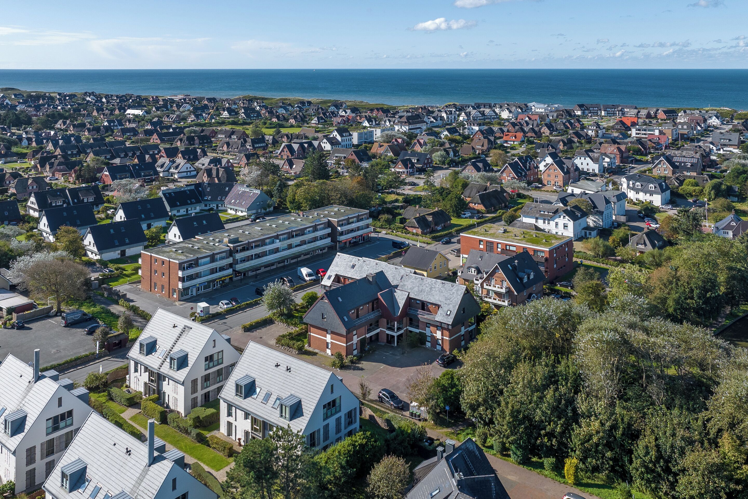 Ett flygbildsbild av ett bostadsområde med hus, en stor byggnad med ett platt tak och en vattenyta i bakgrunden.