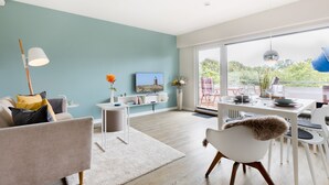 A modern living room with a teal wall, a flat-screen TV, a dining area with a table and chairs, and a large window offering a view of greenery. - Design Kleinod für Preisbewusste, in Bester, Zentraler Lage (Wenningstedt-Braderup (Sylt))