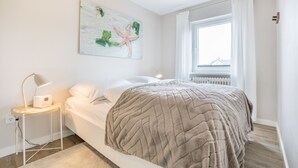 A bedroom with a bed, a bedside table with a lamp, a window with curtains, and a wall-mounted artwork. - Design Kleinod für Preisbewusste, in Bester, Zentraler Lage (Wenningstedt-Braderup (Sylt))