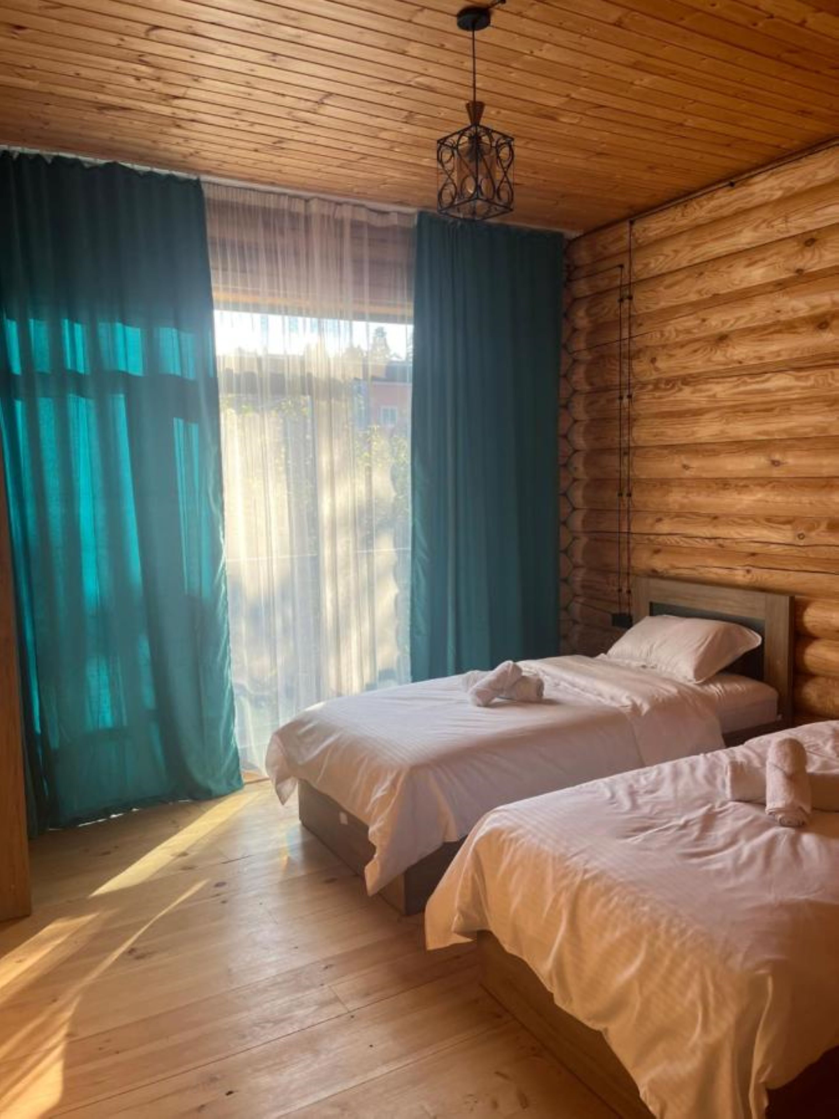 A room with wooden walls and ceiling, two beds with white linens, a hanging light fixture, and a view through a window with teal curtains.