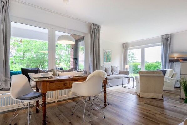 A modern living room with a wooden dining table, white chairs, a sofa, and a large window offering a view of greenery. - Idyllische Lage, Ebenerdig im Erdgeschoss mit Terrasse und Garten (Wenningstedt-Braderup (Sylt))