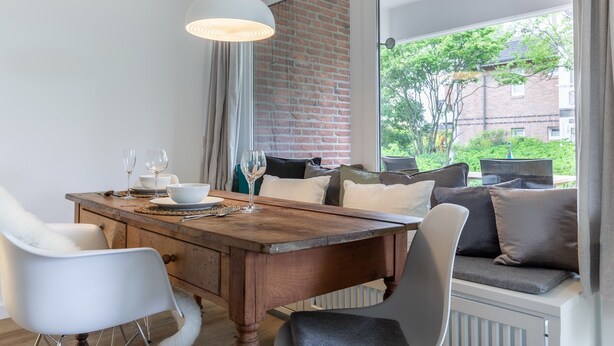 A dining area with a wooden table, white chairs, and a view of a brick building and greenery outside. - Idyllische Lage, Ebenerdig im Erdgeschoss mit Terrasse und Garten (Wenningstedt-Braderup (Sylt))