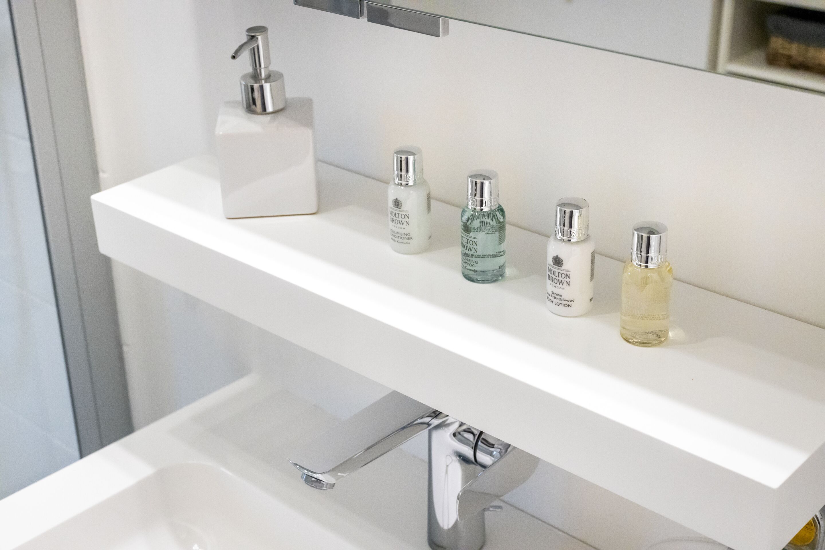 Une salle de bain avec un lavabo blanc, un robinet argenté et quatre flacons de lotion sur l’étagère.