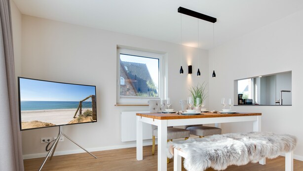 Ein moderner Essbereich mit einem Holztisch, einer weißen Bank und einem Fernseher, der eine Strandansicht zeigt.