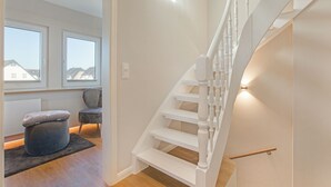 Eine moderne Treppe mit weißen Geländern und einer Holzhandläufe, die zu einem Raum mit einem grauen Pouf und einem Sessel führt.