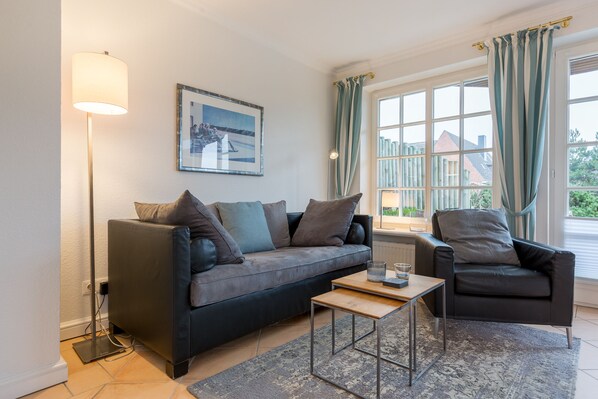 A modern living room with a sofa, armchair, and coffee table, a floor lamp, and a large window with curtains. - Sylt. Heide. Und Meer (Wenningstedt-Braderup (Sylt))